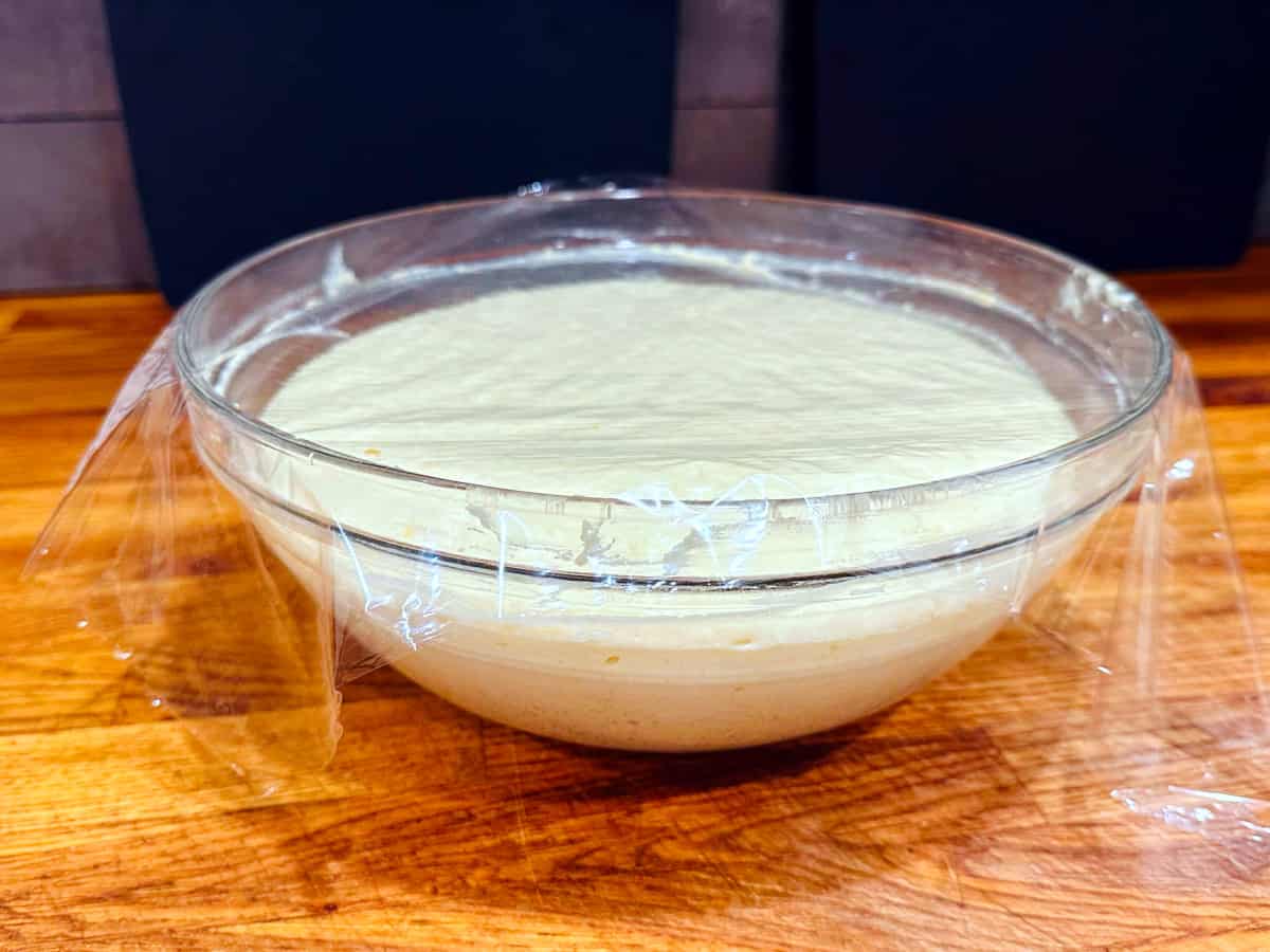 Homemade Belgian waffle batter rising in a large glass bowl covered loosely with plastic wrap.