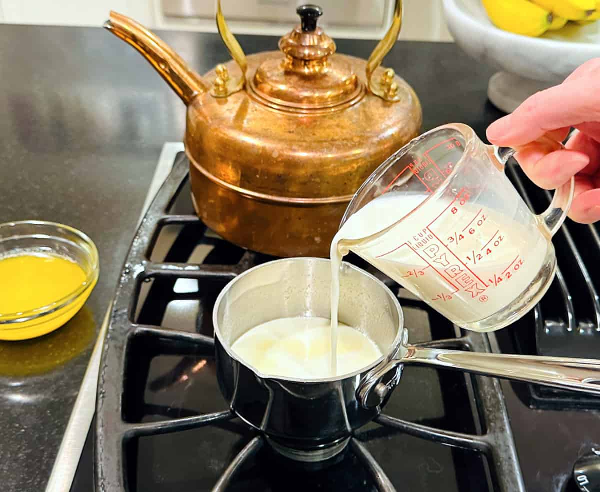 Milk being poured from a glass measuring cup into a small steel saucepan on the stove.