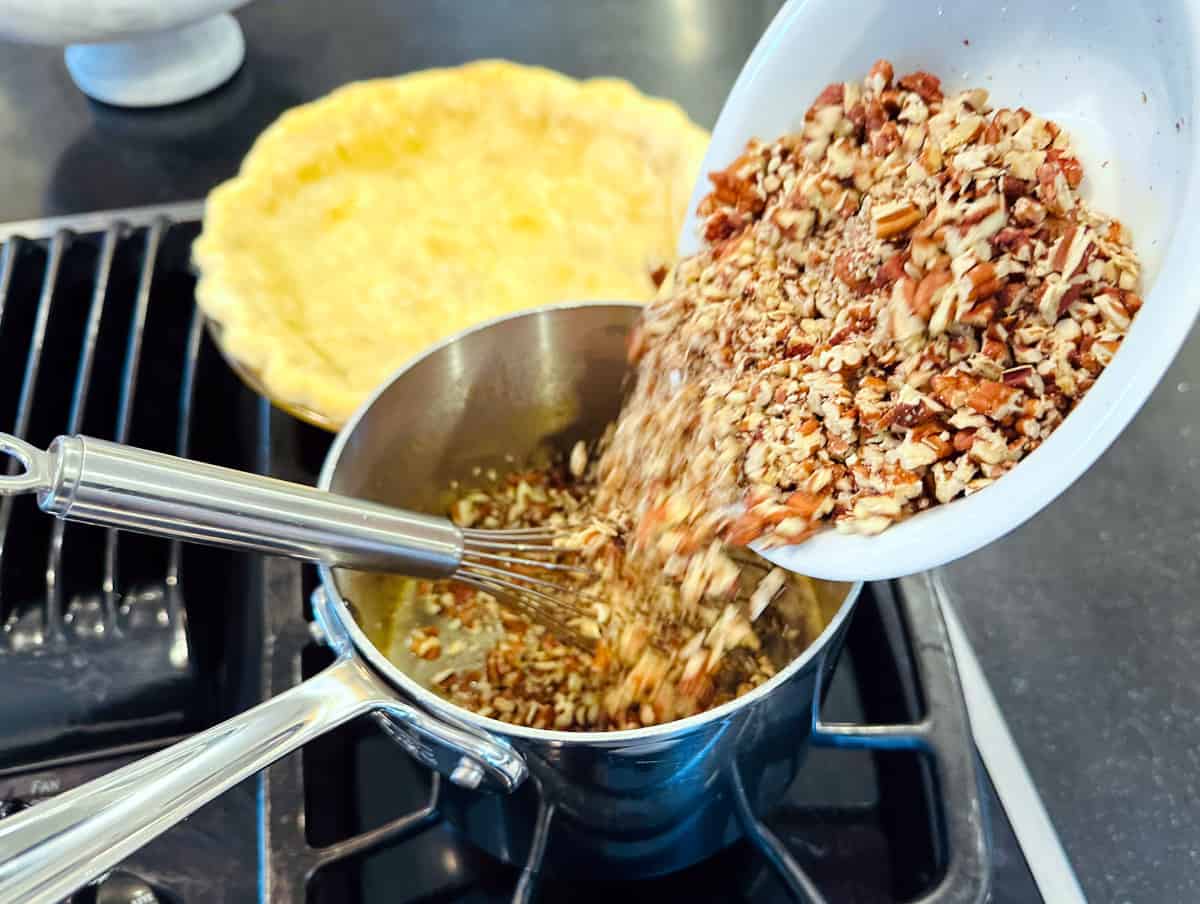 Chopped pecans being tipped from a white bowl into a steel saucepan containing brown liquid and a whisk.