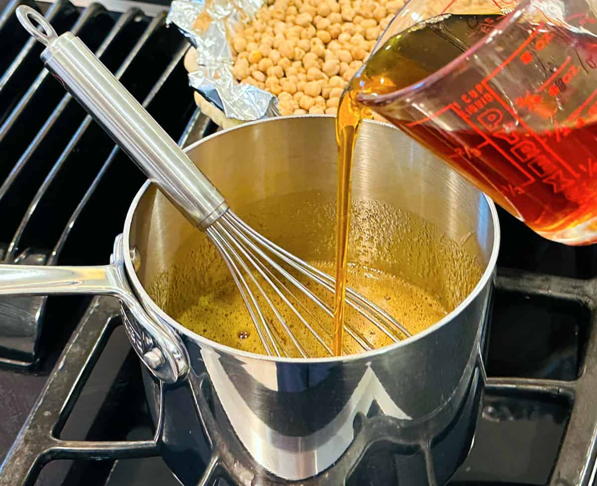 Maple syrup being poured from a glass measuring cup into a steel saucepan containing brown liquid and a metal whisk.