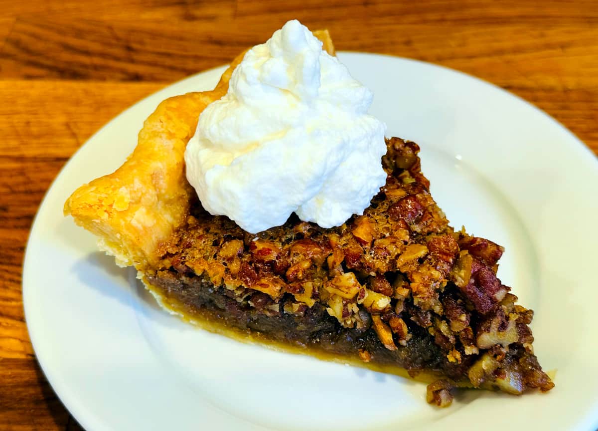 Slice of pecan pie on a small white plate with a big dollop of whipped cream on top.