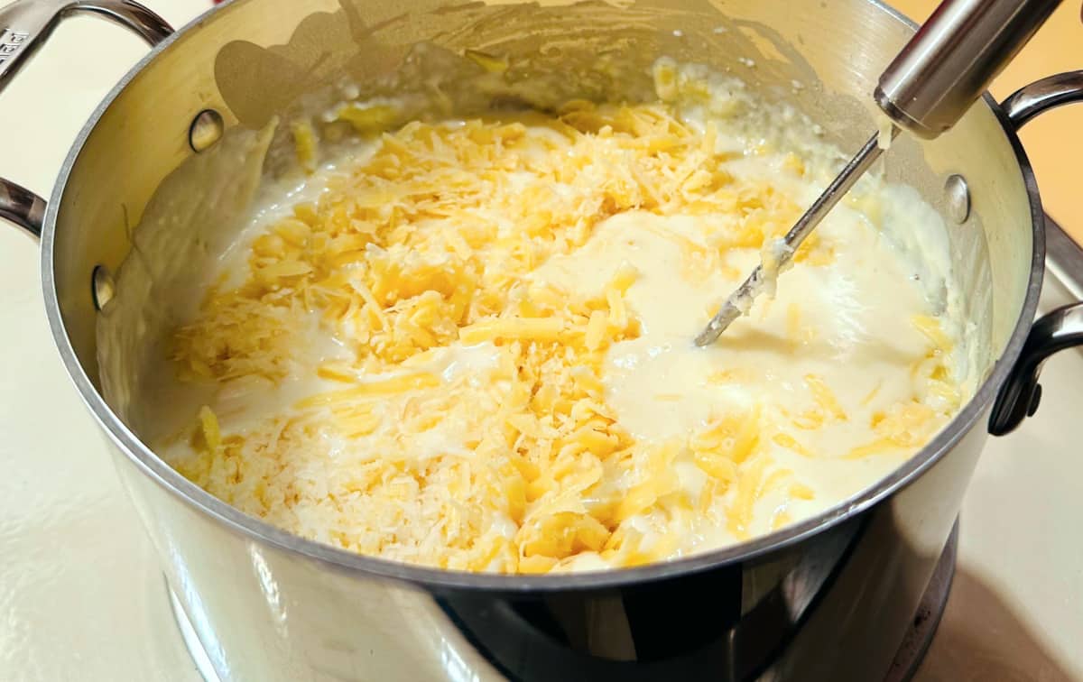 Shredded cheese being mixed into a white creamy mixture of milk and roux in a steel pot.