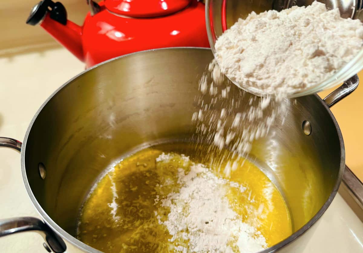 Flour being tipped from a glass bowl into melted butter in a steel pot.