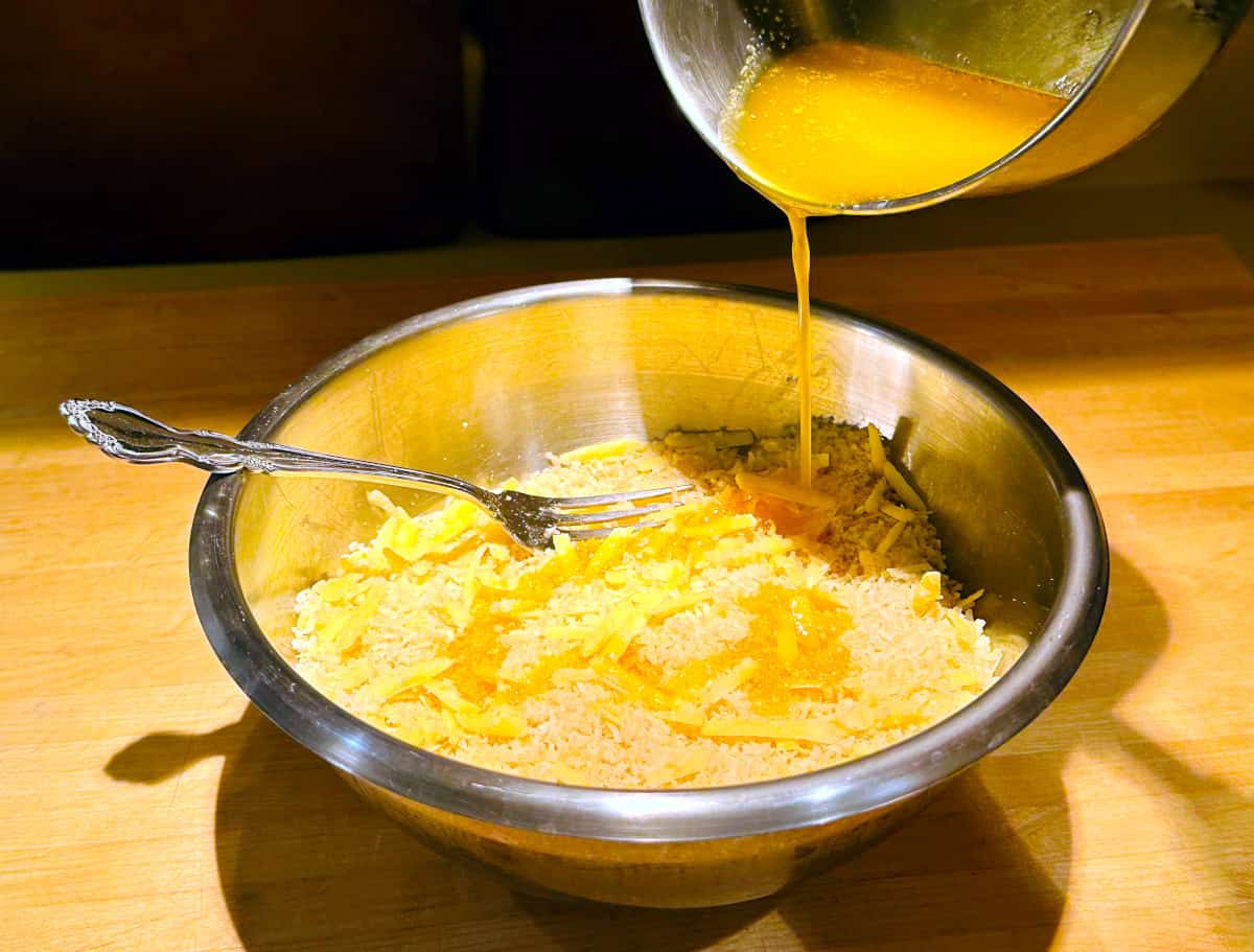 Melted butter being poured from a small steel saucepan into panko and shredded cheese in a steel bowl with a fancy steel fork.