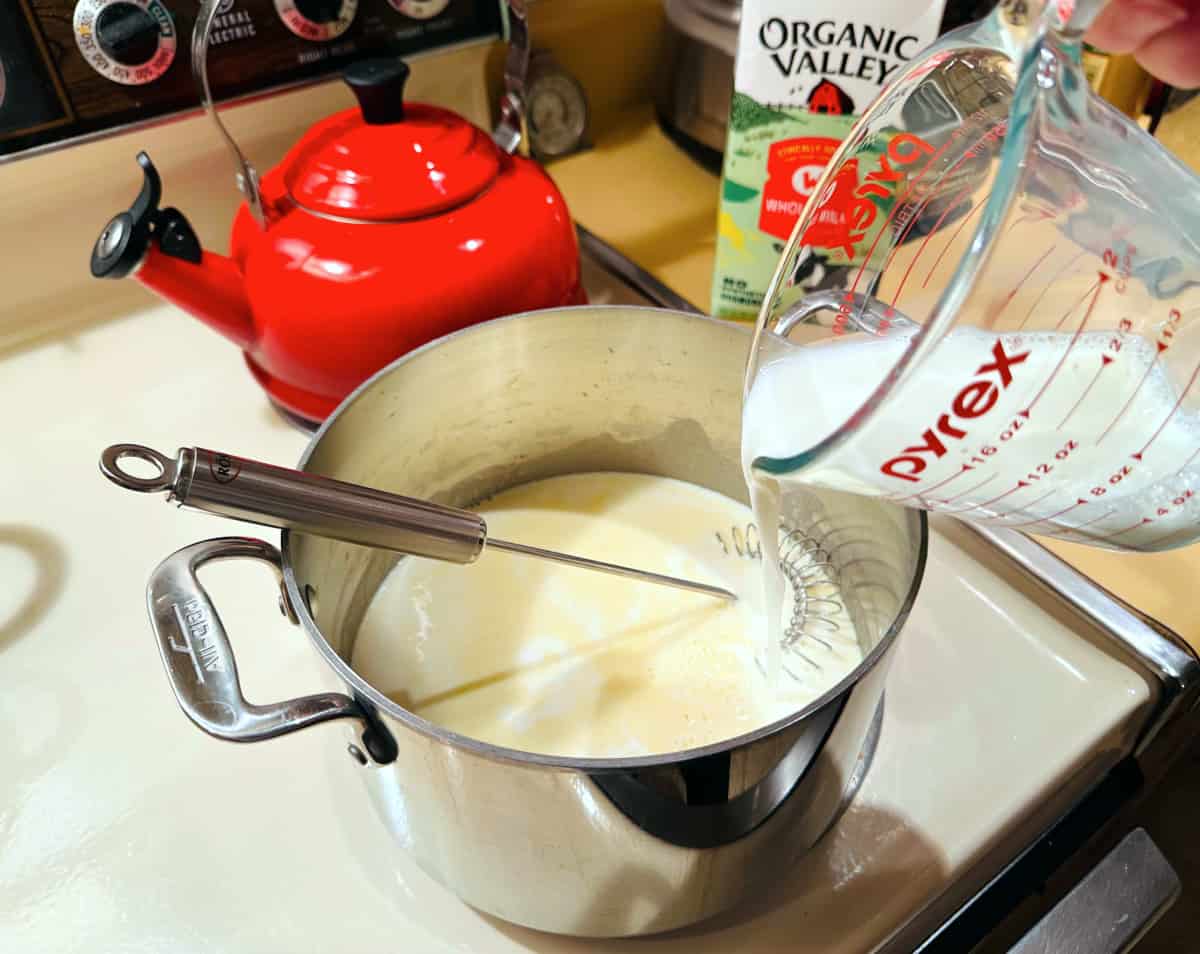 Milk being poured from a glass measuring cup into a steel pot with a sauce whisk in it.