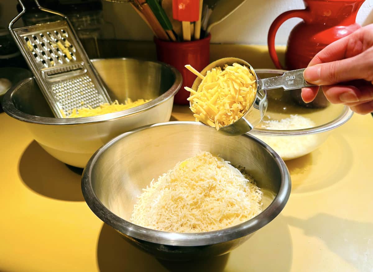 Shredded white cheddar being tipped from a steel measuring cup into panko and shredded Parmigiano Reggiano in a steel bowl.