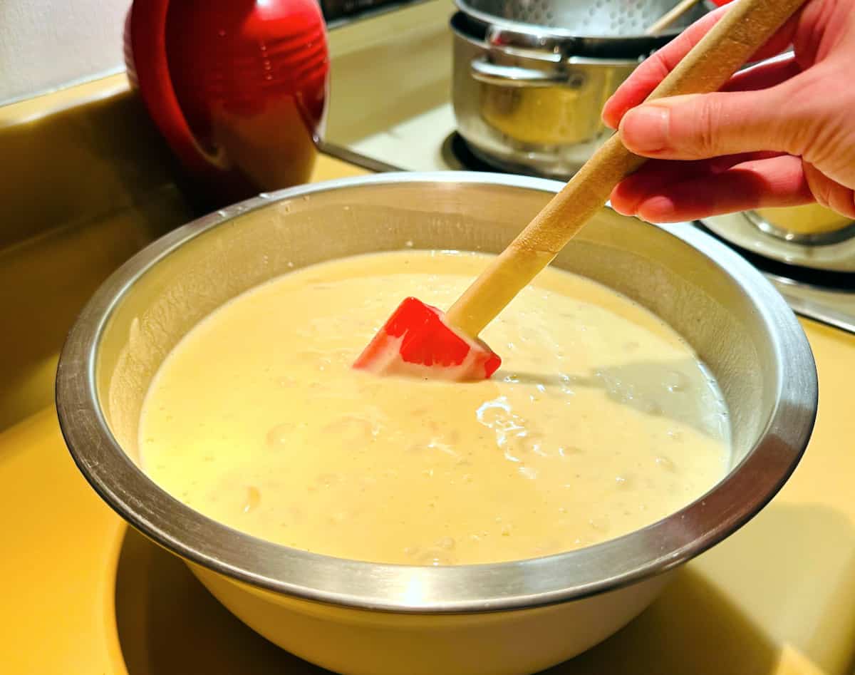 Cooked pasta and cheese sauce being mixed together with a red silicone spatula in a steel bowl.