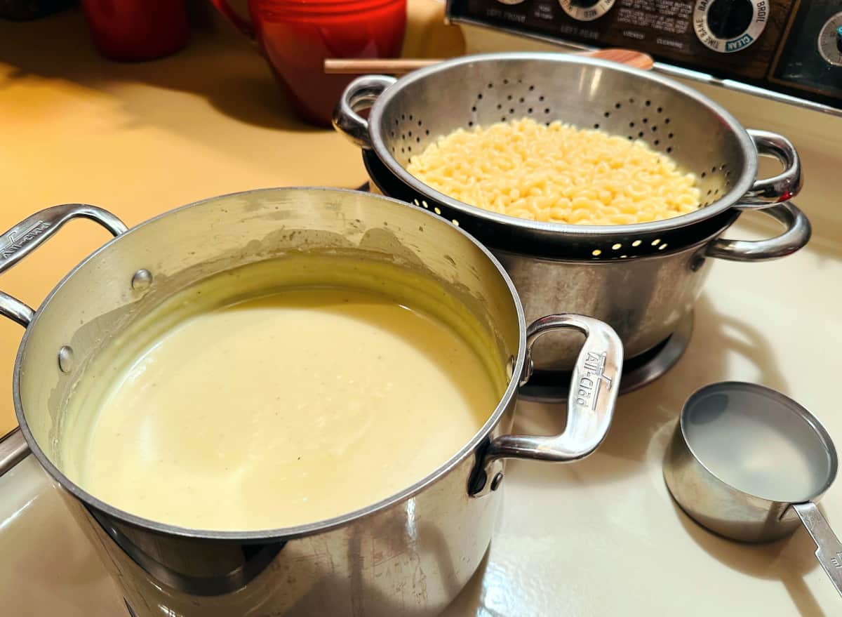 Cheese sauce mixture in a steel pot sitting next to a colander full of cooked macaroni noodles and a steel measuring cup full of reserved pasta cooking water.