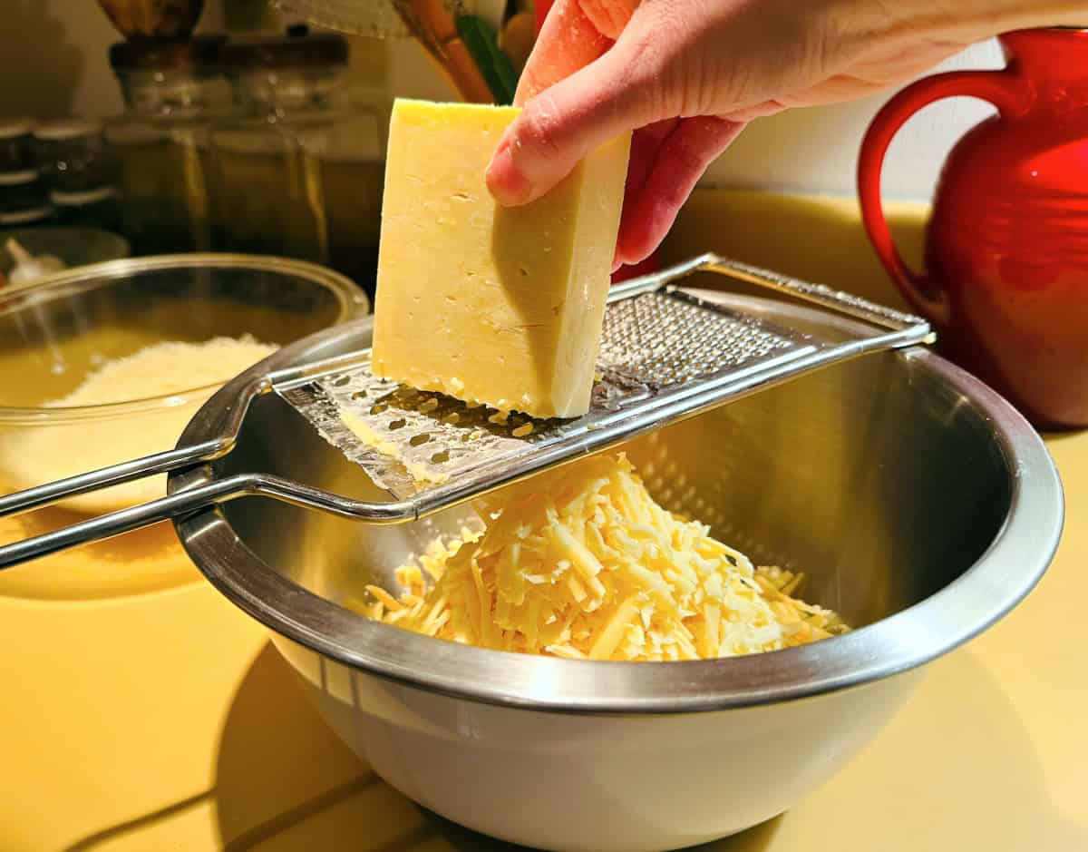 Block of white cheddar being coarsely grated into a large steel bowl.