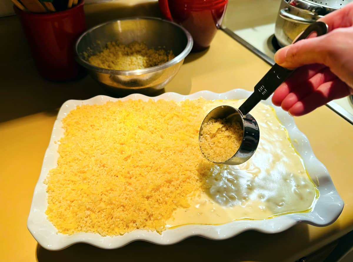 Topping mixture being sprinkled from a steel measuring cup over macaroni and cheese mixture in a white baking dish with a ruffled rim.