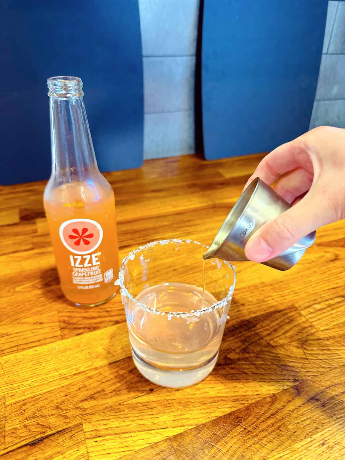 Pale pink liquid being poured from a steel measuring jigger into an old fashioned glass with a salted rim.