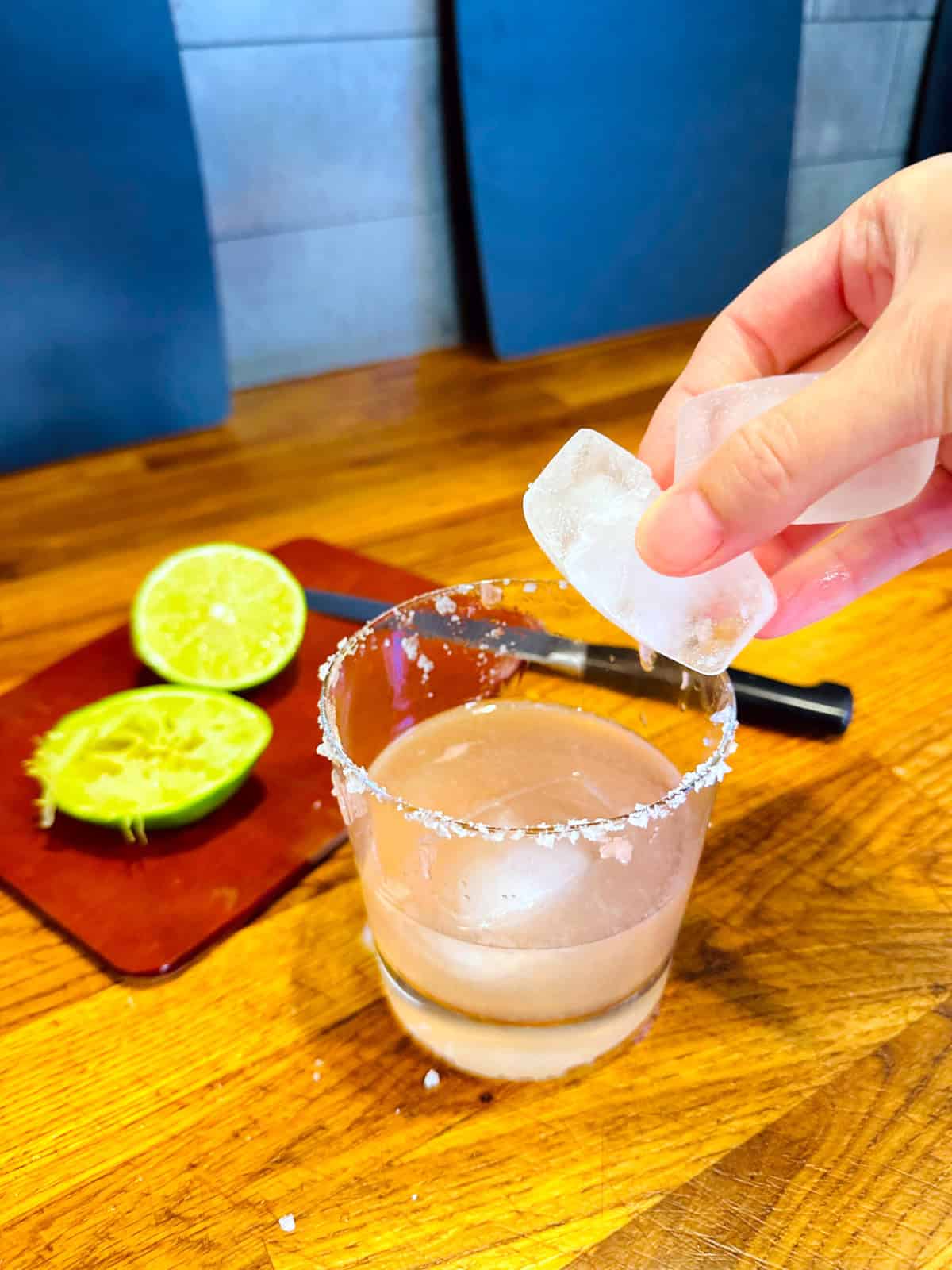 Chunks of ice being dropped into a glass with a salted rim containing pale pink liquid.