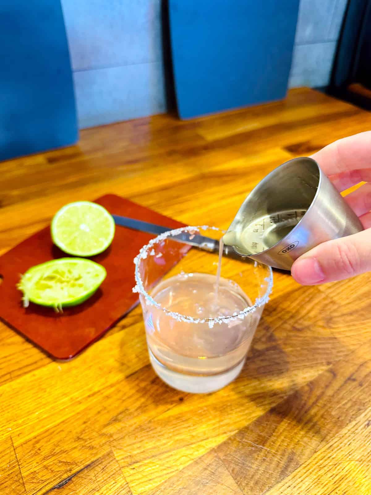 Pale green liquid being poured from a steel measuring jigger into an old fashioned glass with a salted rim next to two halves of lime on a small brown cutting board.