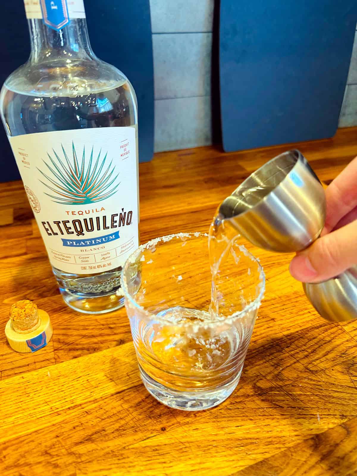 Clear liquid being poured from a steel jigger into an old fashioned glass with a salted rim next to a bottle of tequila.