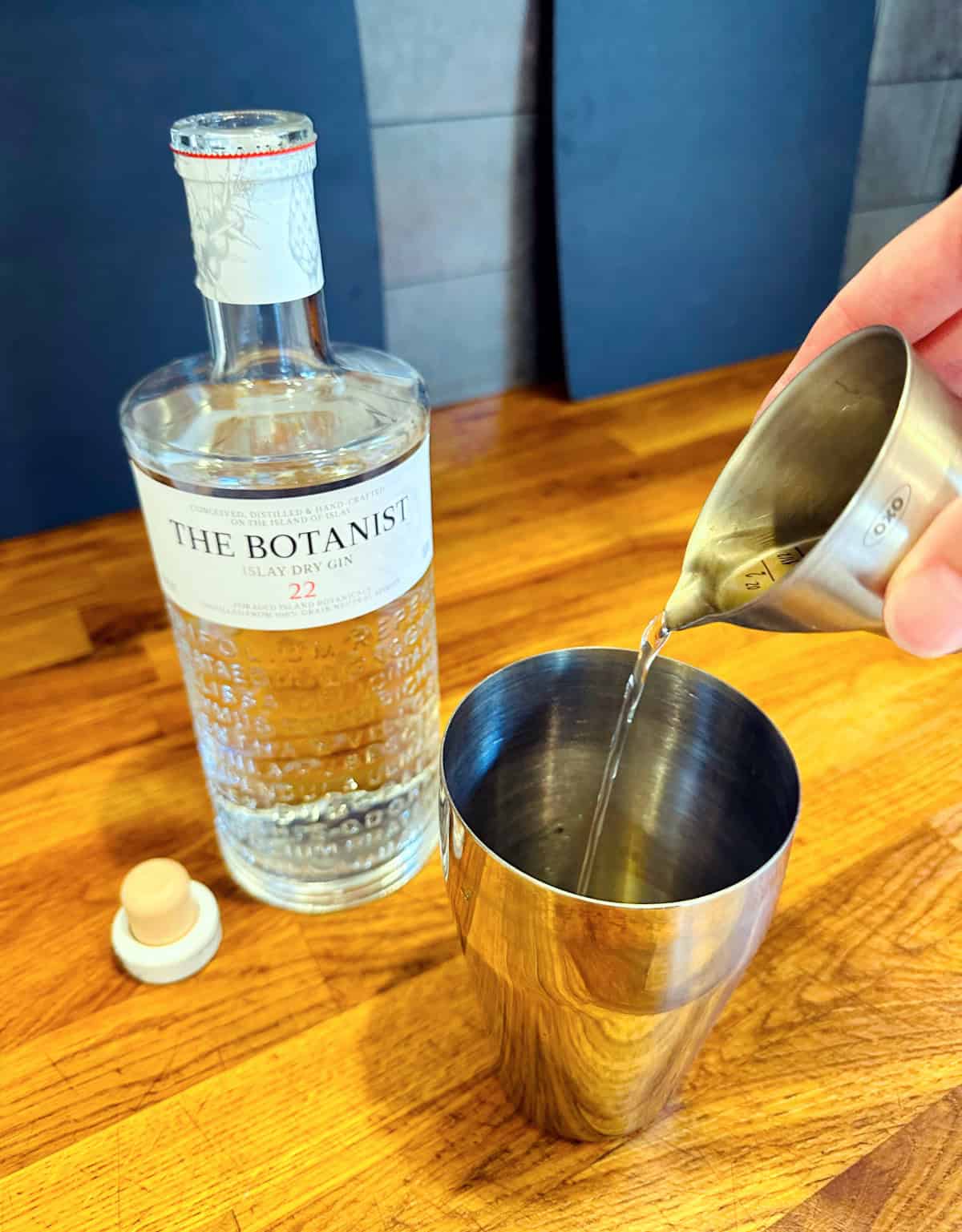 Clear liquid being poured from a steel measuring jigger into a steel cocktail shaker next to a bottle of gin.