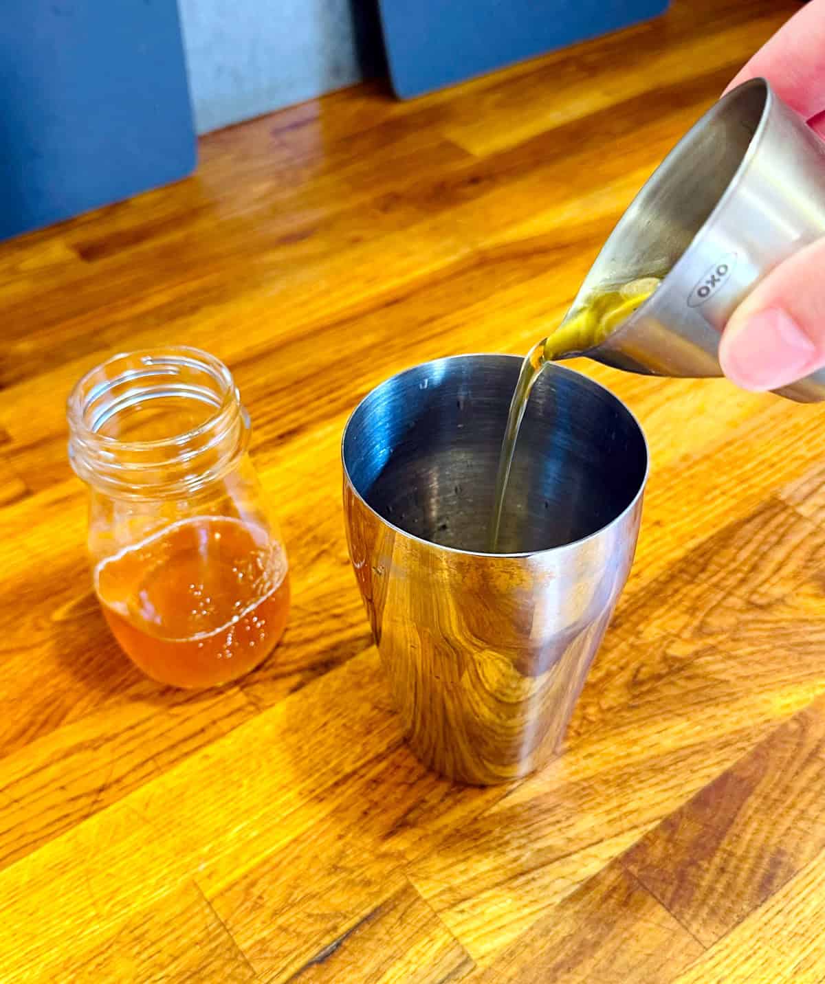 Pale golden liquid being poured from a steel measuring jigger into a steel cocktail shaker next to a glass jar containing pale golden liquid.