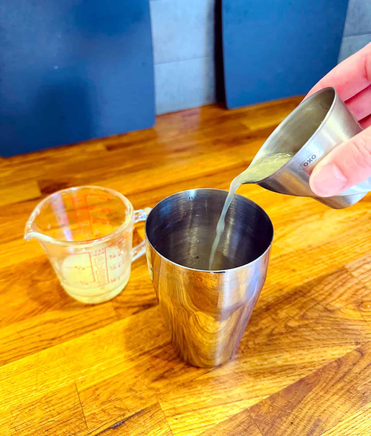 Yellow liquid being poured from a steel measuring jigger into a steel cocktail shaker next to a glass measuring cup containing yellow liquid.