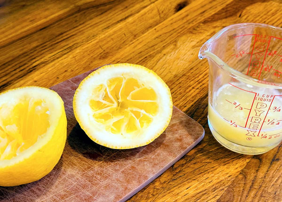 Two lemon halves on a brown cutting board next to a glass measuring cup containing lemon juice.