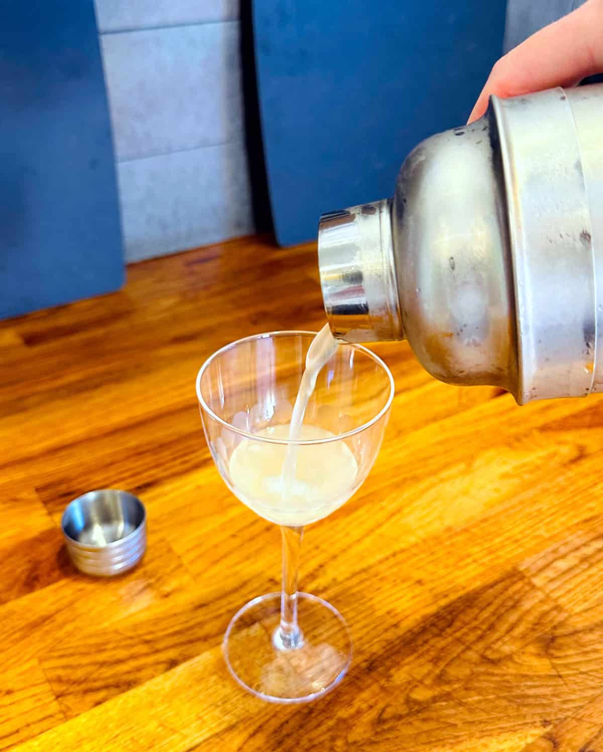 Pale yellow liquid being poured from a frosty steel cocktail shaker into a coupe glass.