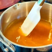 Golden yellow liquid being stirred with a small white silicone spatula in a small steel saucepan.
