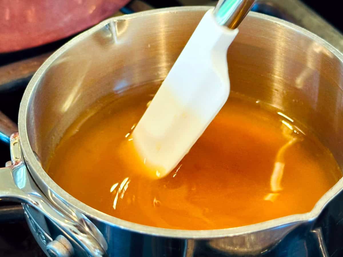 Golden yellow liquid being stirred with a small white silicone spatula in a small steel saucepan.