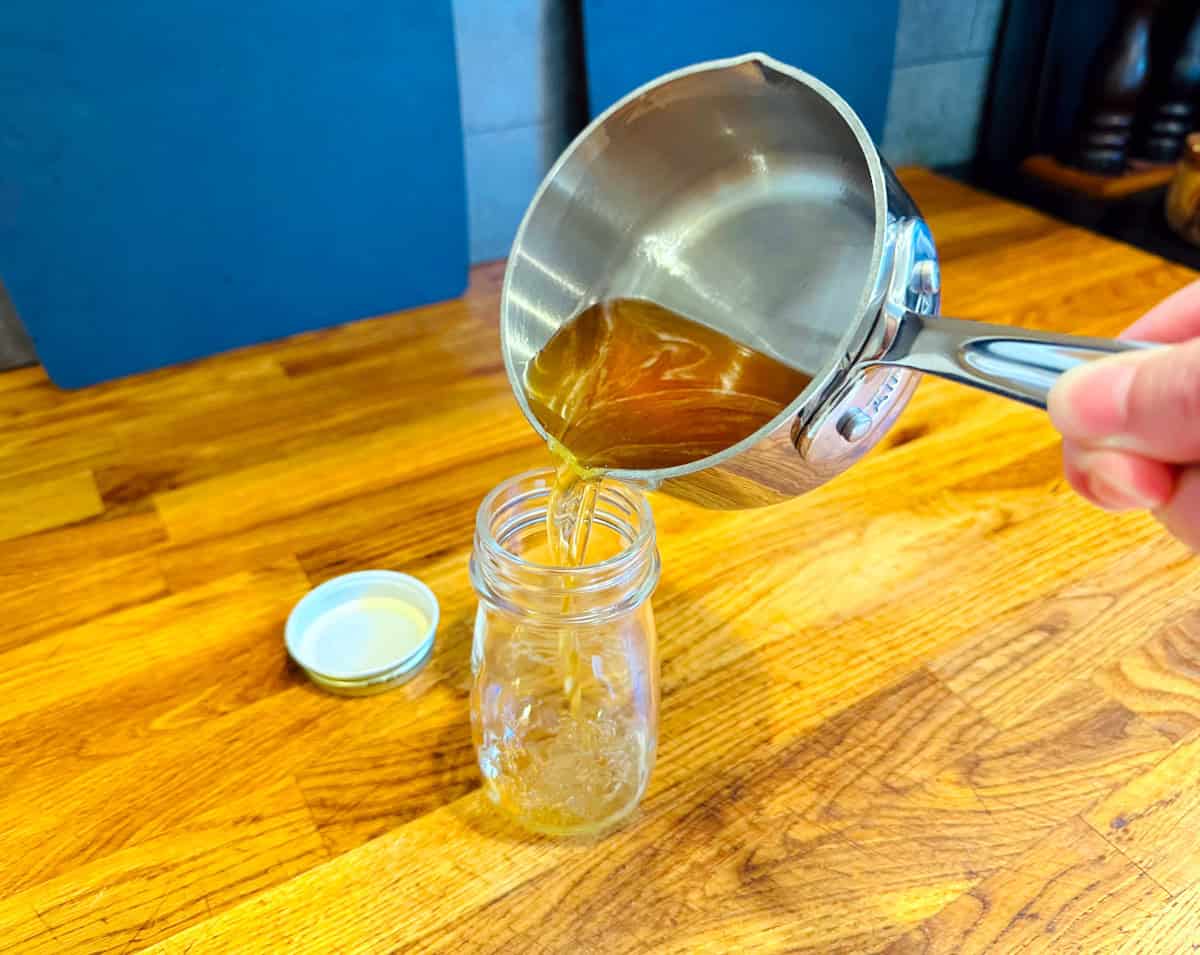Golden yellow liquid being poured from a small steel saucepan into a glass jar.