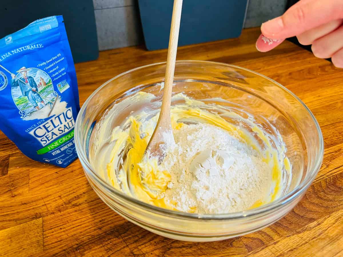 Pinch of sea salt being added to a large glass bowl containing yellow batter with a small heap of flour on top and a wooden spoon.