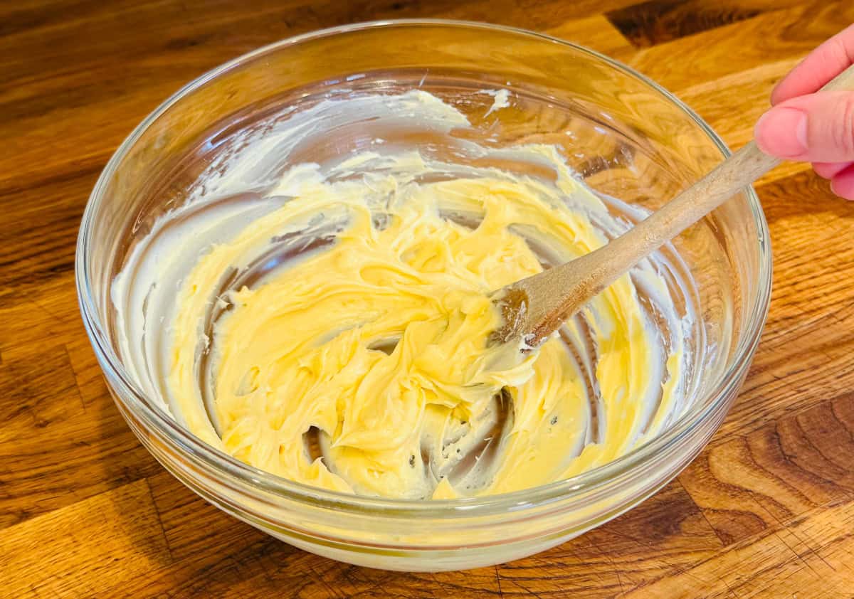 Pale yellow mixture of butter and sugar being beaten with a wooden spoon in a glass bowl.