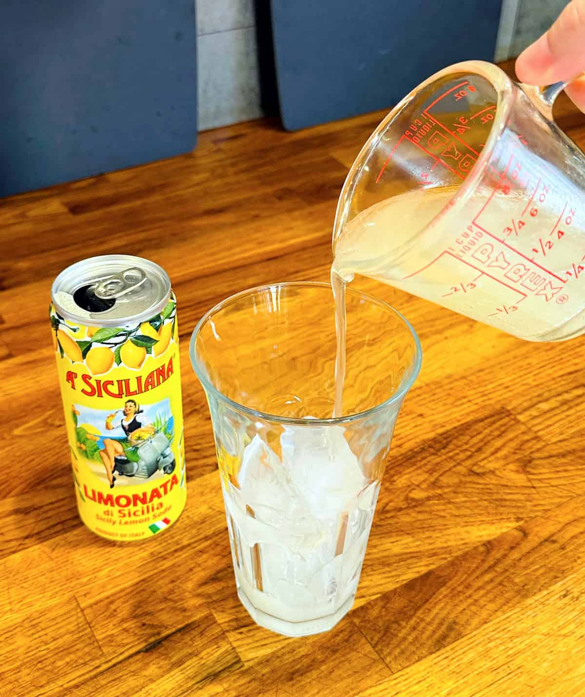 Pale yellow liquid being poured from a glass measuring cup into a tall glass of ice next to a can of lemon soda.