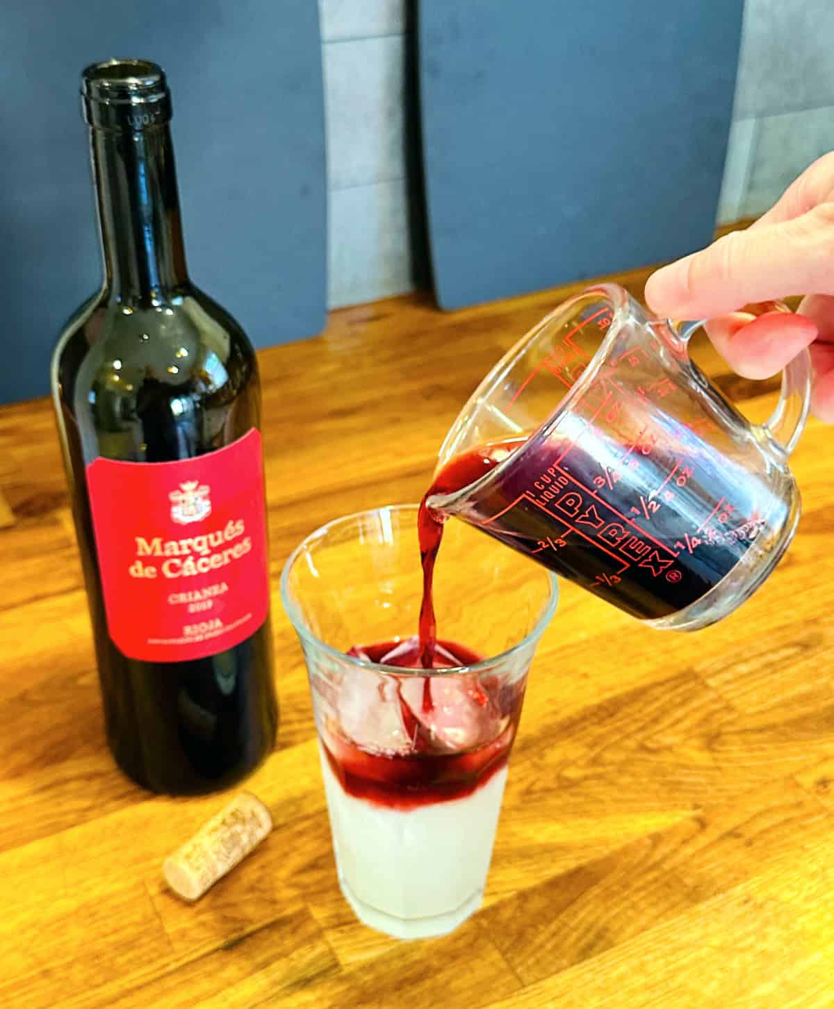 Dark red liquid being poured from a glass measuring cup into a tall glass of pale yellow liquid and ice next to a bottle of red wine.