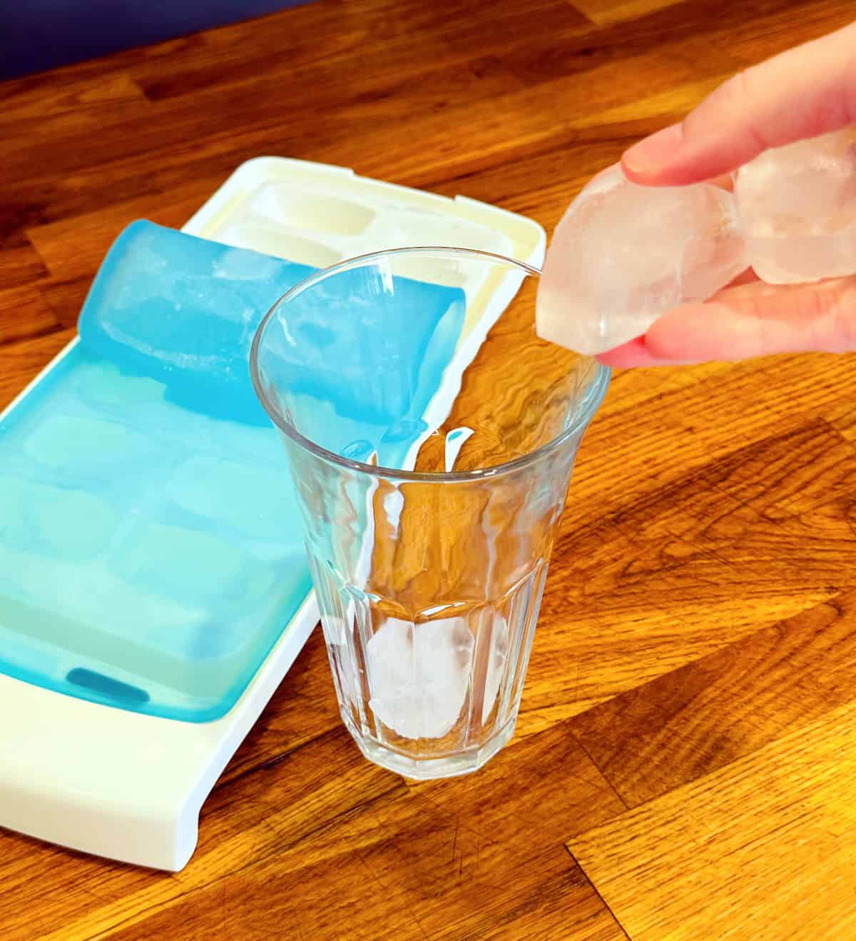 Ice cubes being dropped into a tall glass next to an ice cube tray with a light blue top.