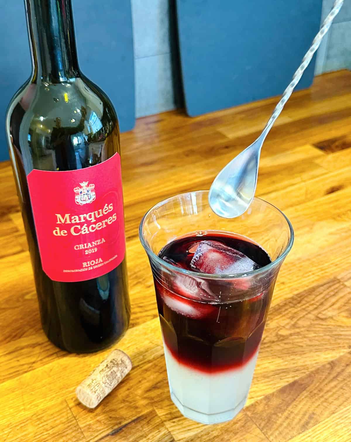 Long steel spoon held over tall glass containing red wine, lemon soda, and ice next to a bottle of wine with a red label and a cork.