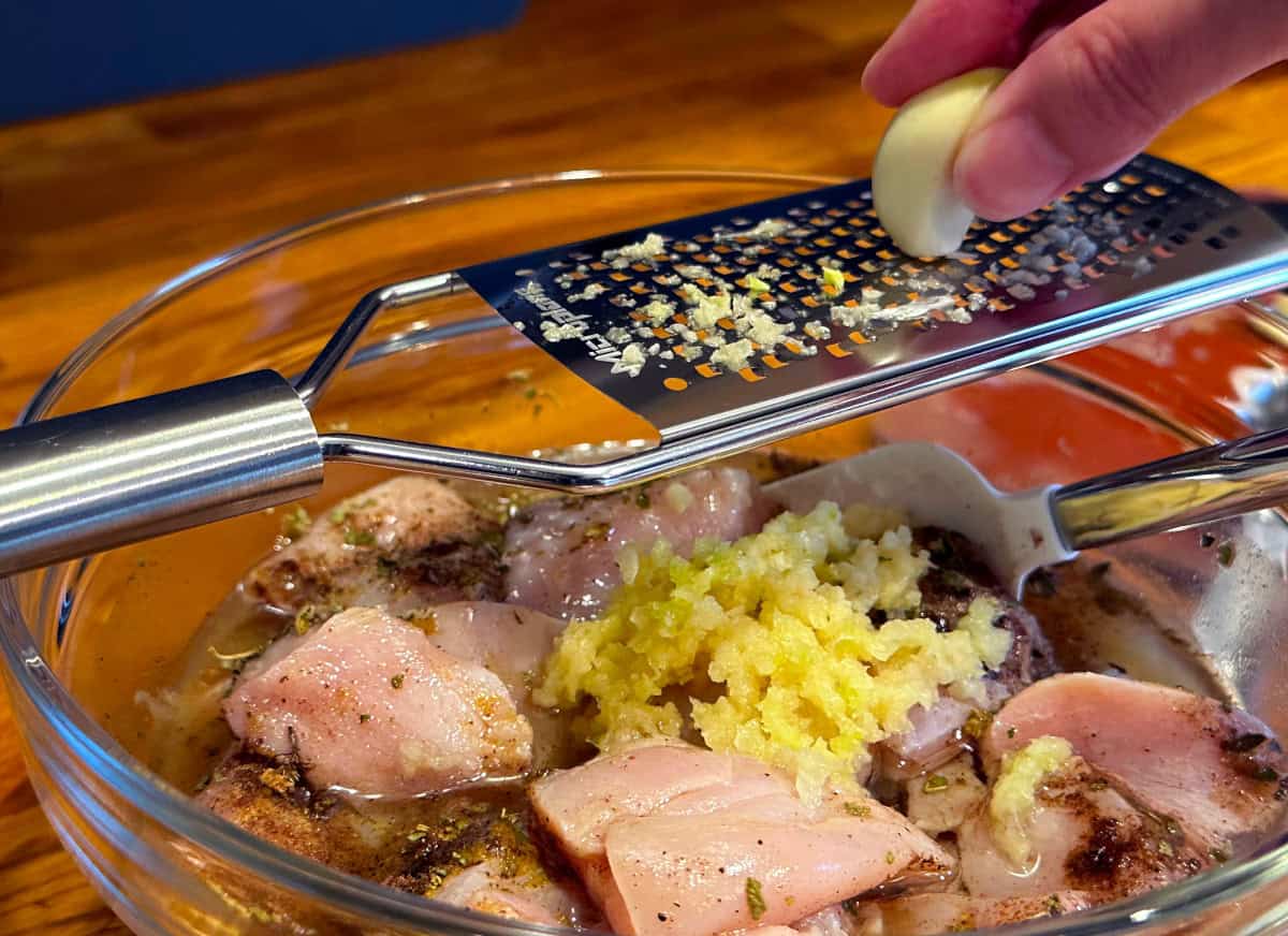 Garlic cloves being grated into a bowl of raw chicken pieces in a citrus marinade.