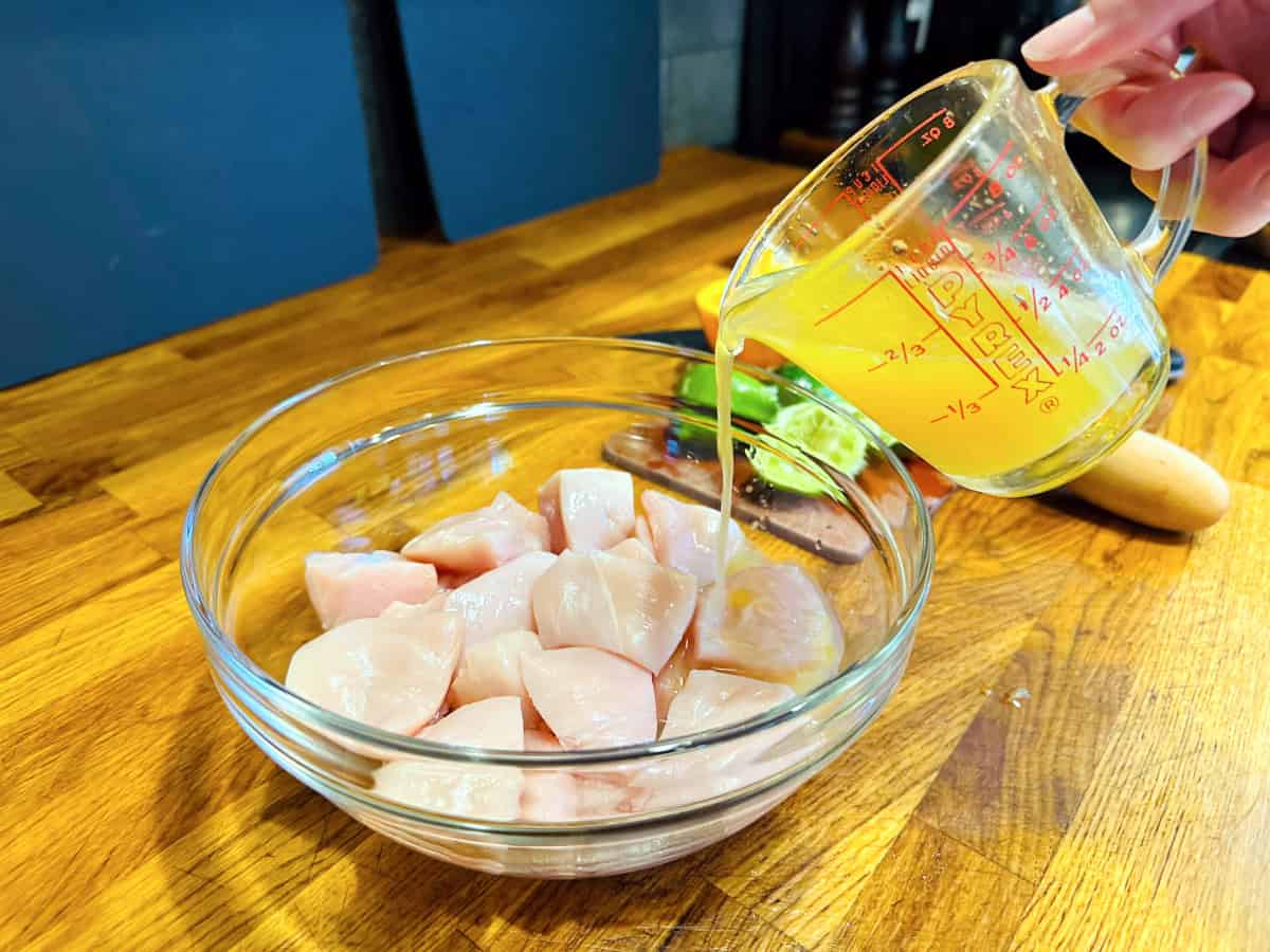 Orange liquid being poured from a glass measuring cup into a glass bowl containing raw chicken pieces.