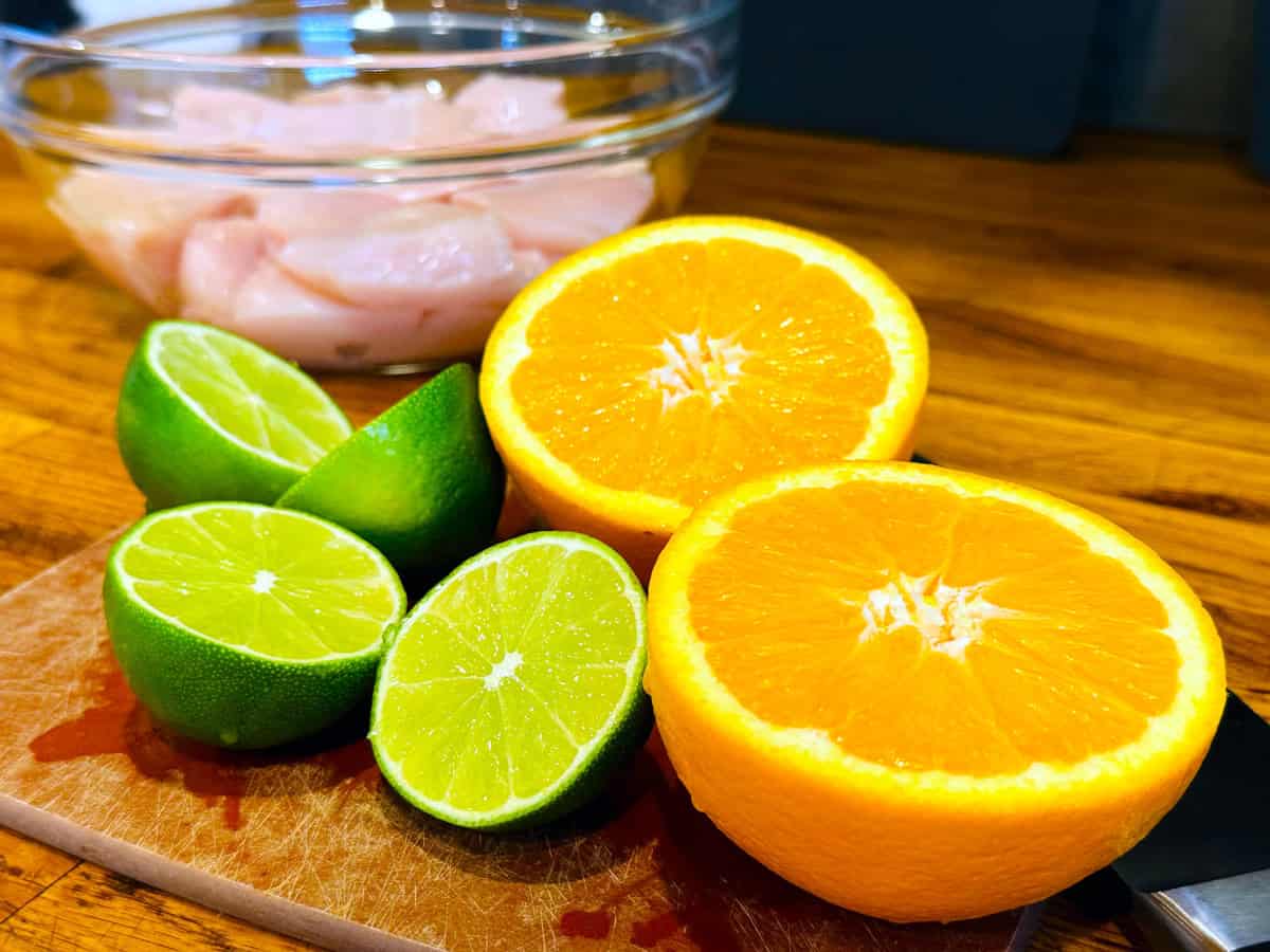 Four lime halves and two orange halves sitting on a cutting board in front of a glass bowl containing raw chicken pieces.