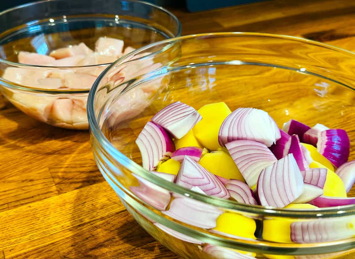 A small glass bowl containing pieces of raw chicken and a medium glass bowl containing chunks of yellow potato and red onion.