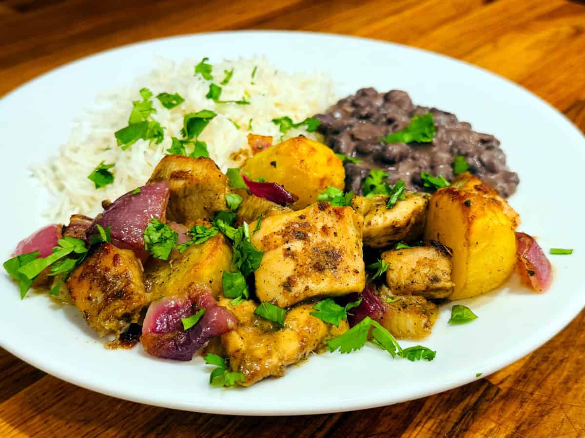 Cuban mojo chicken sprinkled with chopped cilantro and served with a side of black beans and white rice on a white plate.