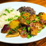 Cuban mojo chicken sprinkled with chopped cilantro and served with a side of black beans and white rice on a white plate.