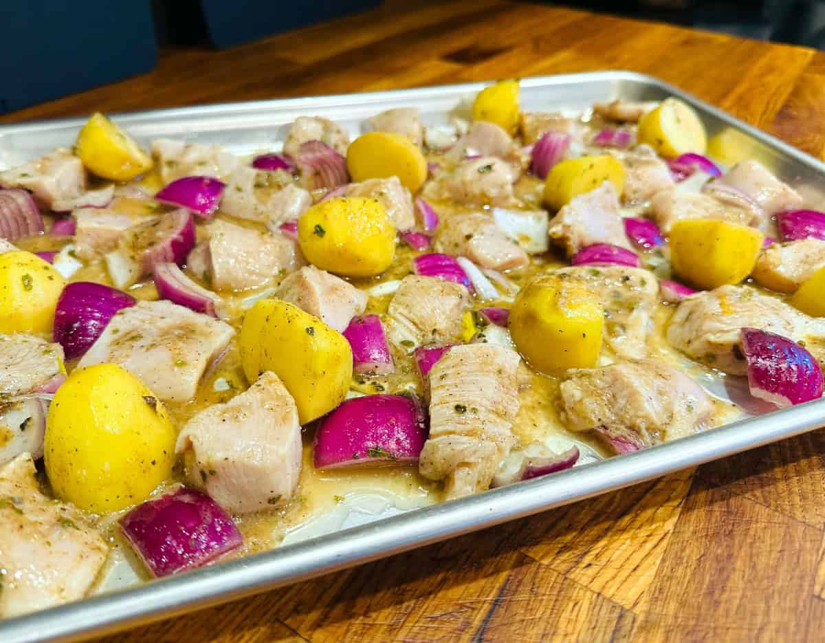 Marinated chicken pieces, yellow potato chunks, and red onion spread out on a metal baking sheet.