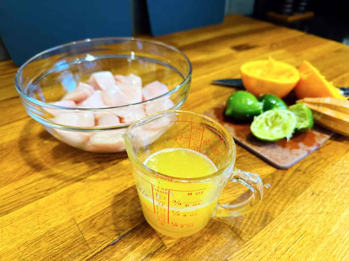 Orange liquid in a glass measuring cup sitting in front of a glass bowl containing raw chicken pieces next to a cutting board strewn with juiced lime and orange halves and a wooden reamer.