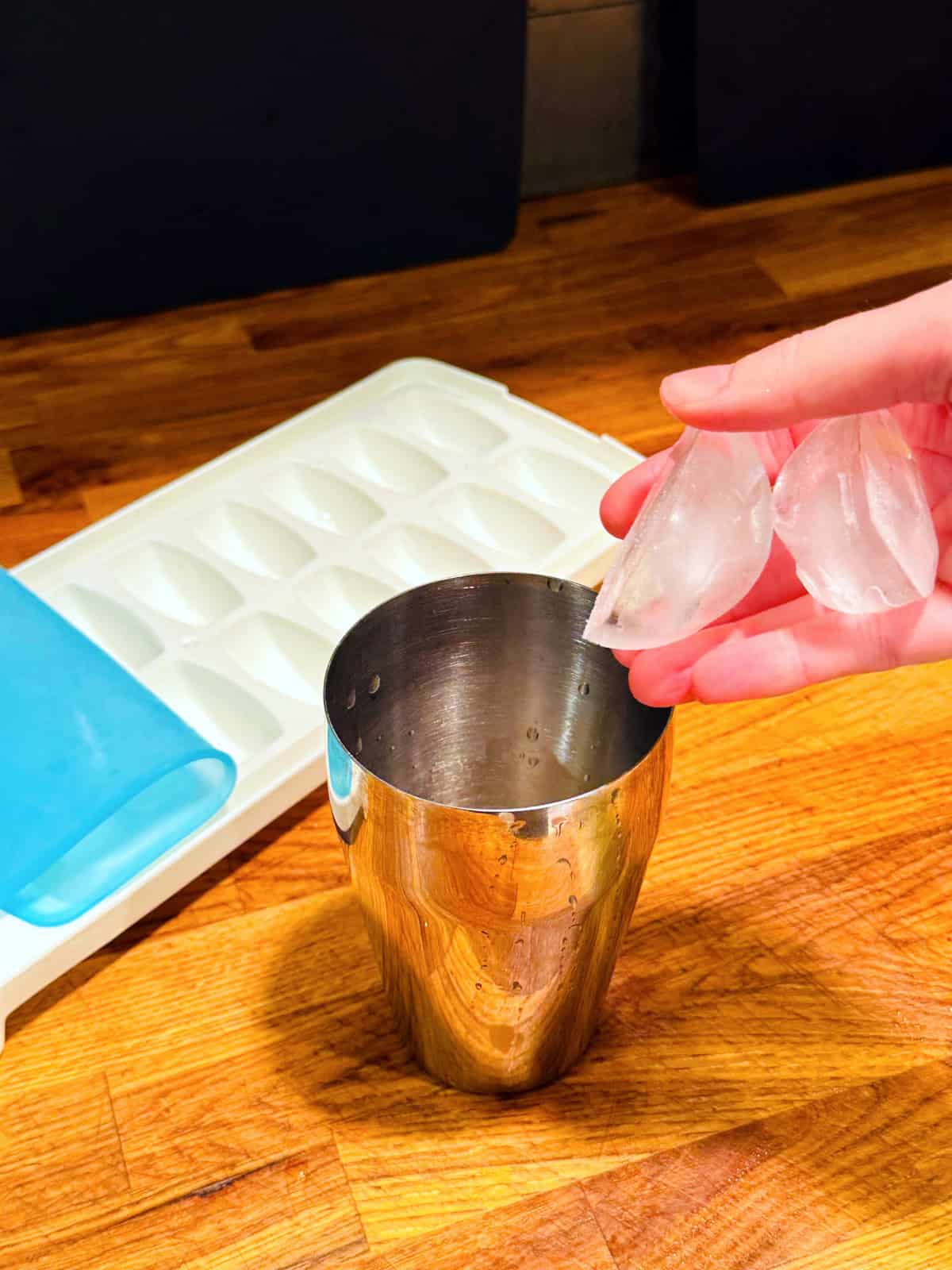 Large ice cubes being dropped into a cocktail shaker sitting next to a white ice cube tray with a light blue lid.