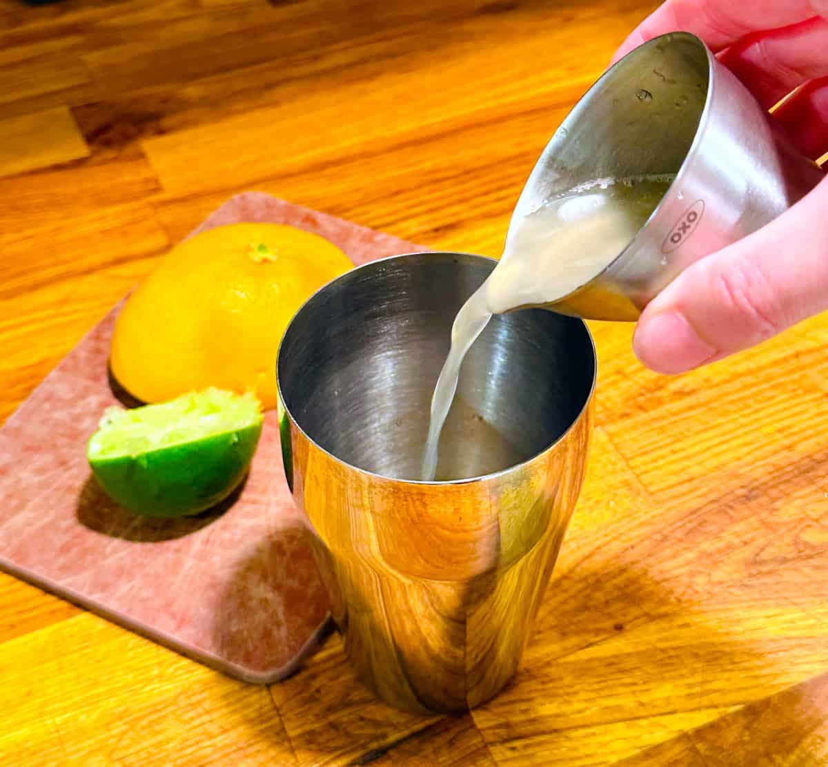 Pale pink liquid being poured from a steel measuring jigger into a cocktail shaker next to the remainder of a lime and a grapefruit half.