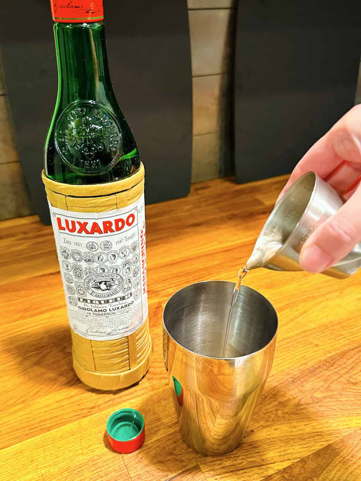 Clear liquid being poured from a steel measuring jigger into a cocktail shaker next to a bottle of maraschino liqueur.