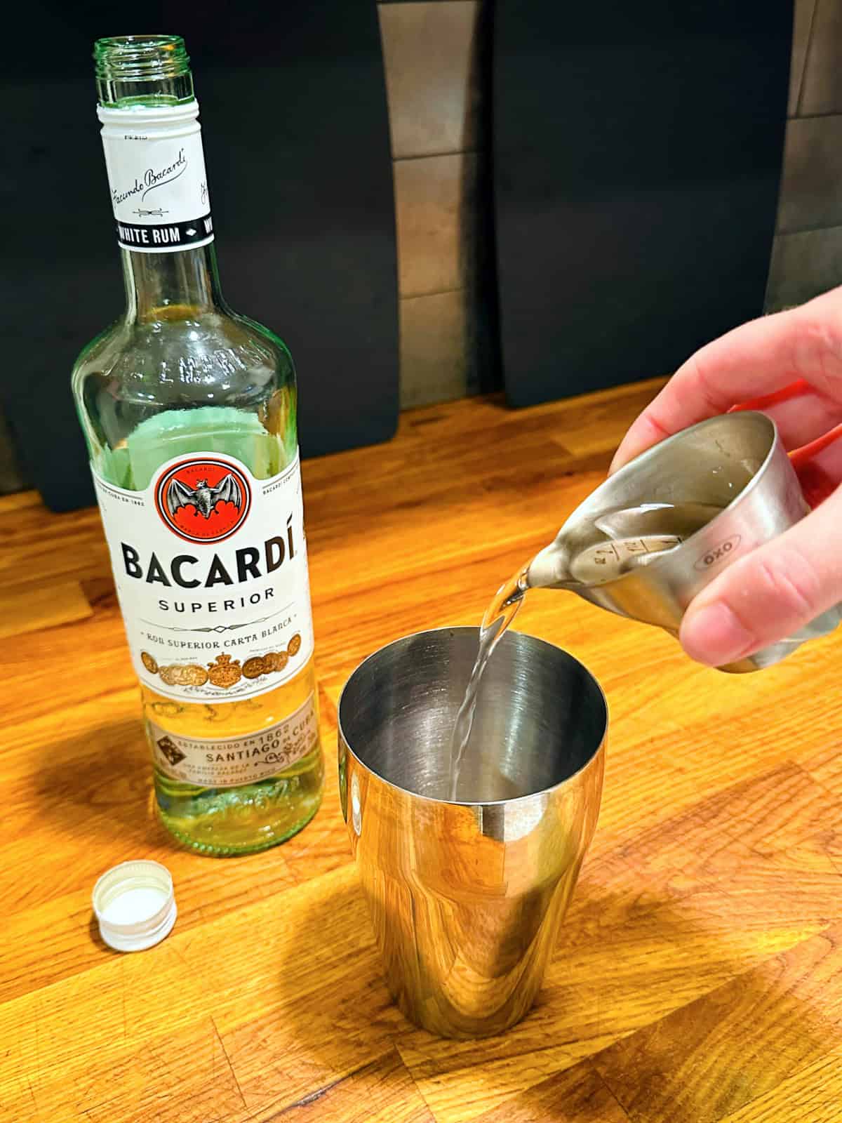 Clear liquid being poured from a steel measuring jigger into a cocktail shaker next to a bottle of white rum.