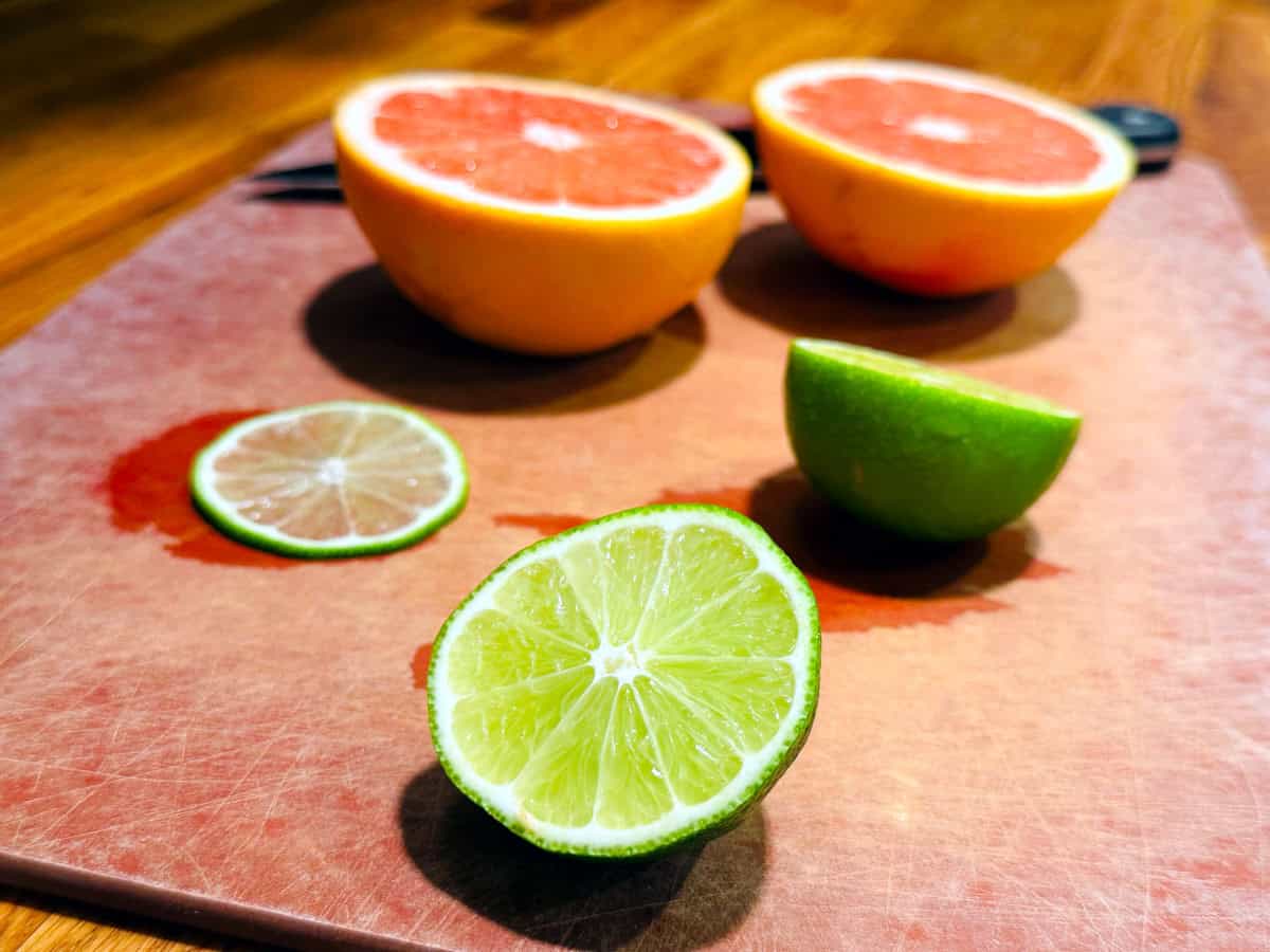 Two halves of lime, a thin slice of lime, and two halves of grapefruit sitting on a cutting board.