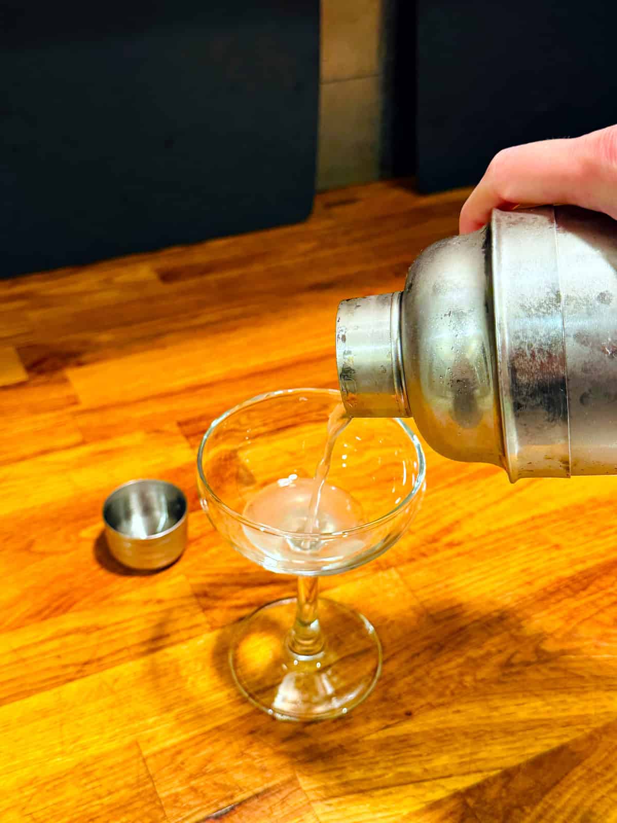 Pale cloudy liquid being poured from a steel cocktail shaker into a coupe glass.