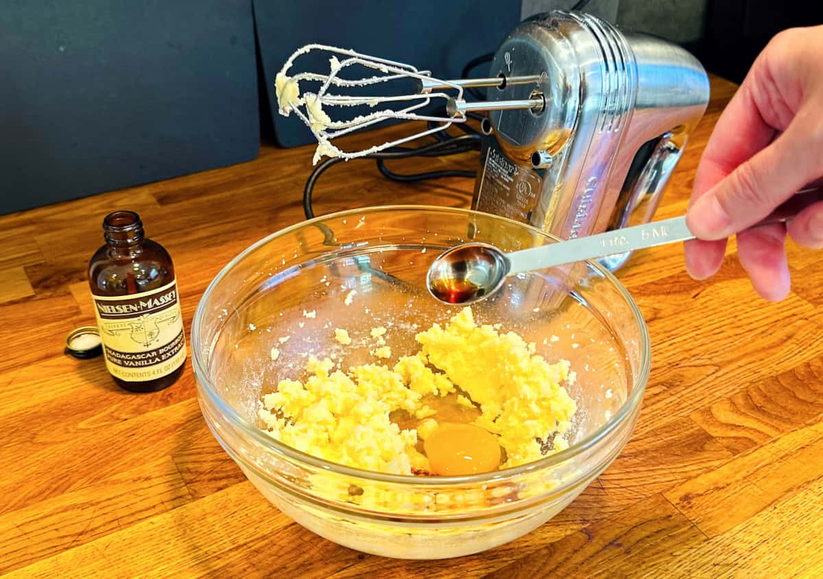 Vanilla extract being tipped from a steel measuring spoon into pale yellow mixture and an egg in a glass bowl.