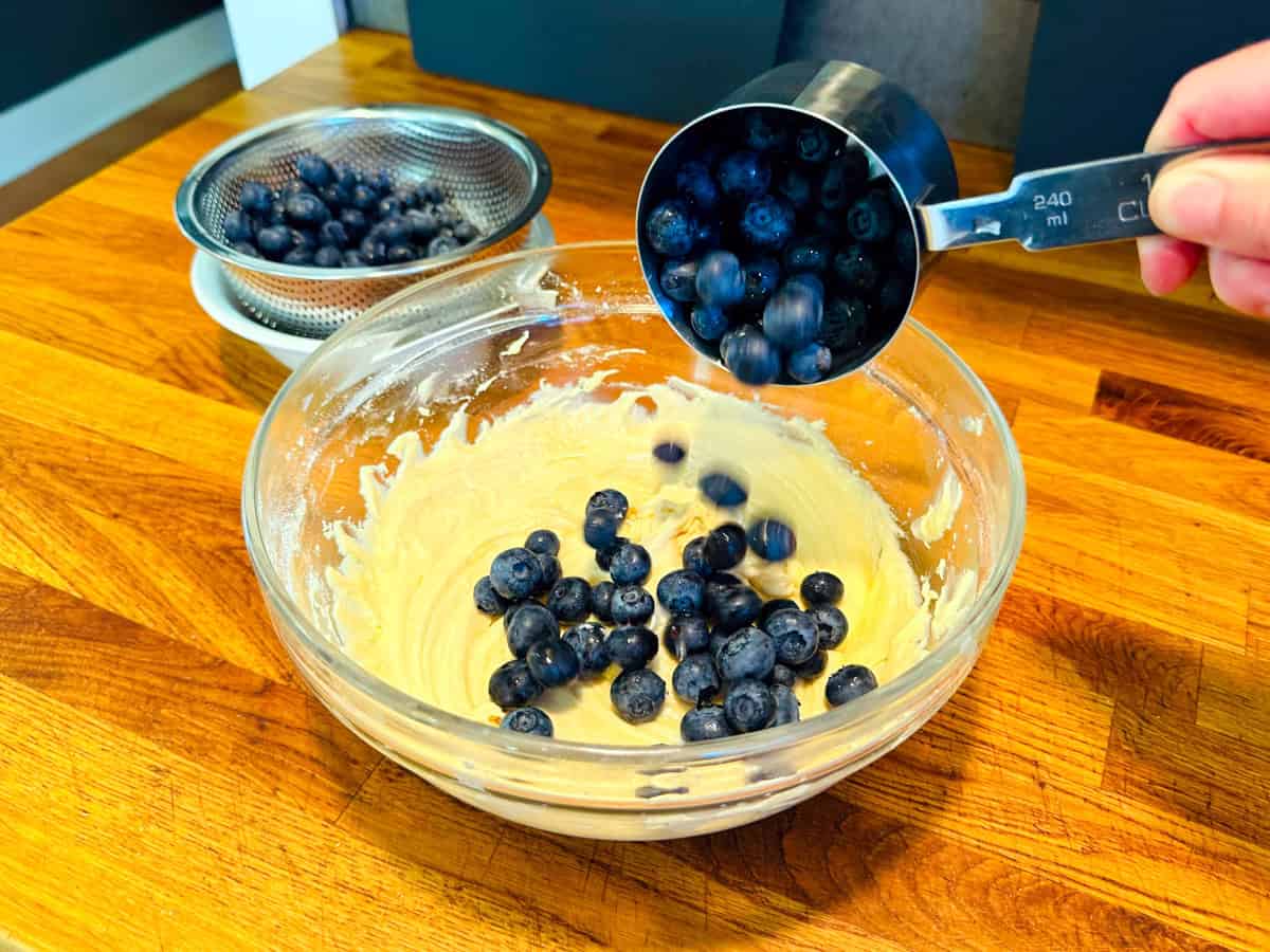 Fresh blueberries being tipped from a steel measuring cup into pale yellow batter in a glass bowl.