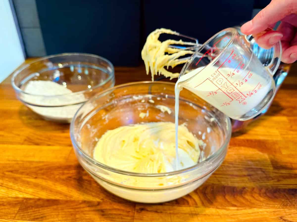 Milk being poured from a glass measuring cup into pale yellow batter in a glass bowl.