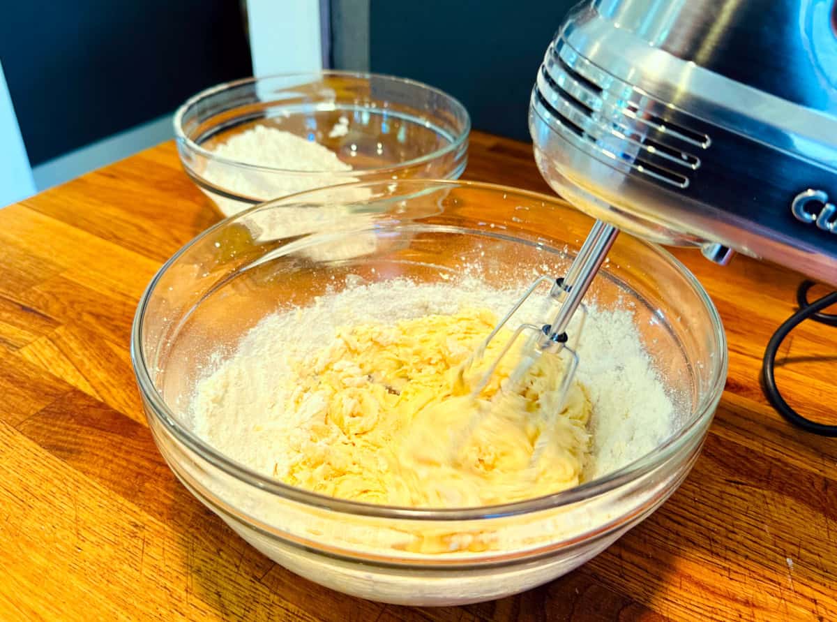 Flour being beaten into butter and sugar mixture with an electric mixer in a glass bowl.